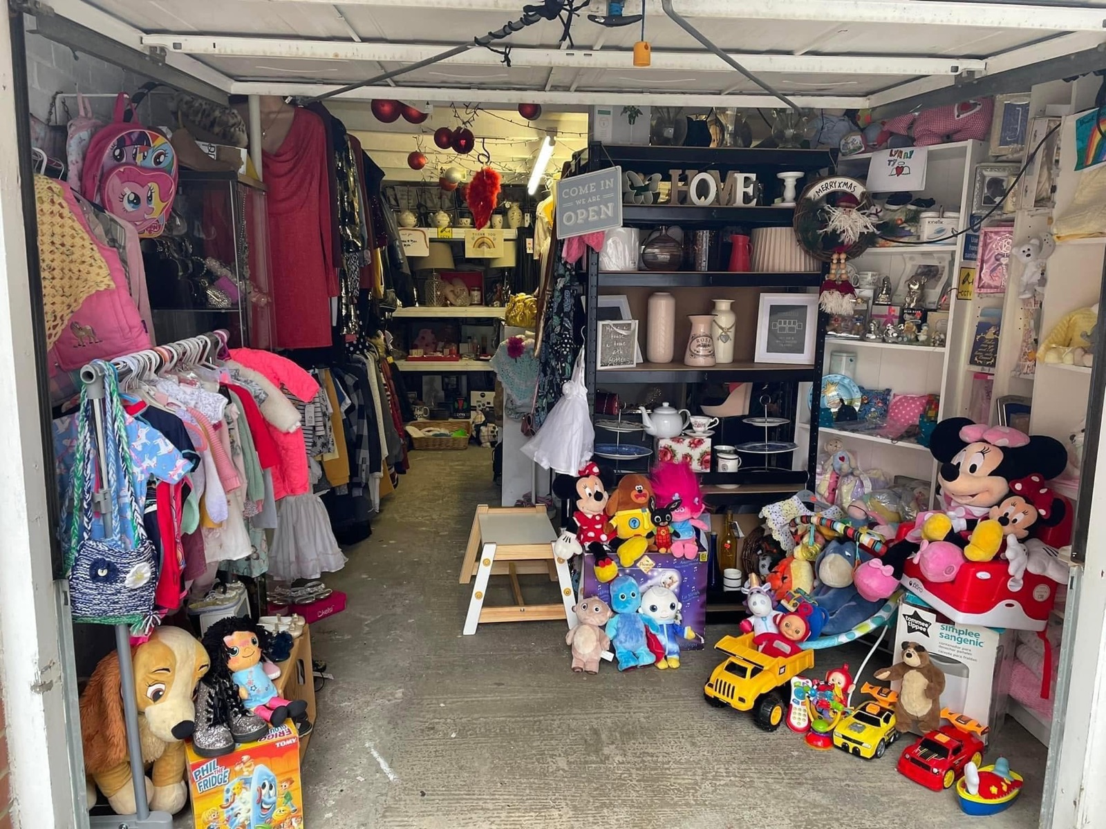 Inside the original Angels Treasure Cove garage, shelves stacked with donated toys, gifts and household items, with a 'come in we are open' sign